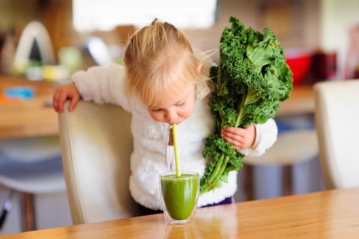 Little toddler girl in a white fluffy jacket holds a bunch of fresh kale and is about to drink a green smoothie in the kitchen