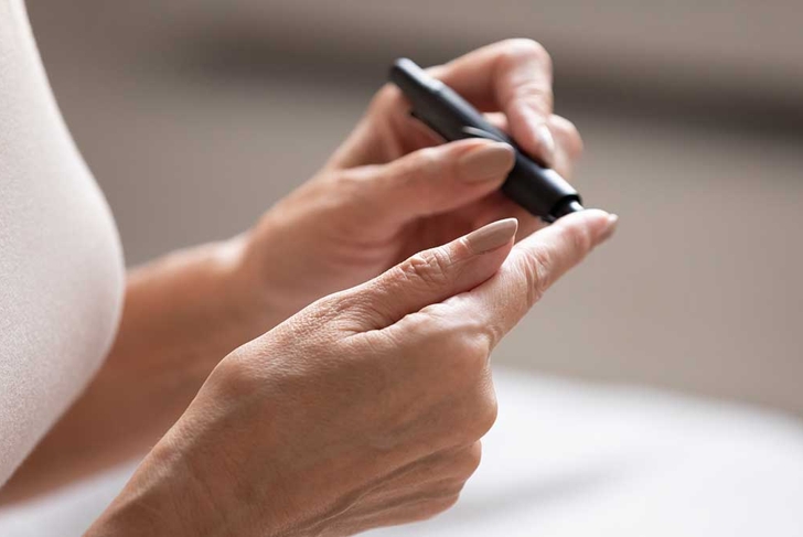 Close up older retired female hands with lancet pen on finger. Mature woman measuring sugar range or glucose level in blood herself in morning at home. Diabetes routine, healthcare domestic concept.